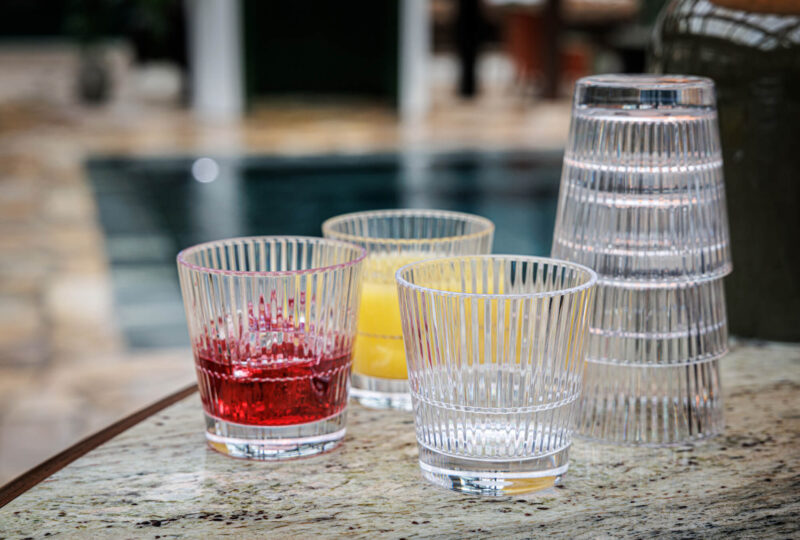 Unbreakable plastic Stripe 355R glasses - red cocktail, yellow drink, and clear striped tumblers on textured surface, showcasing versatile and durable drinkware